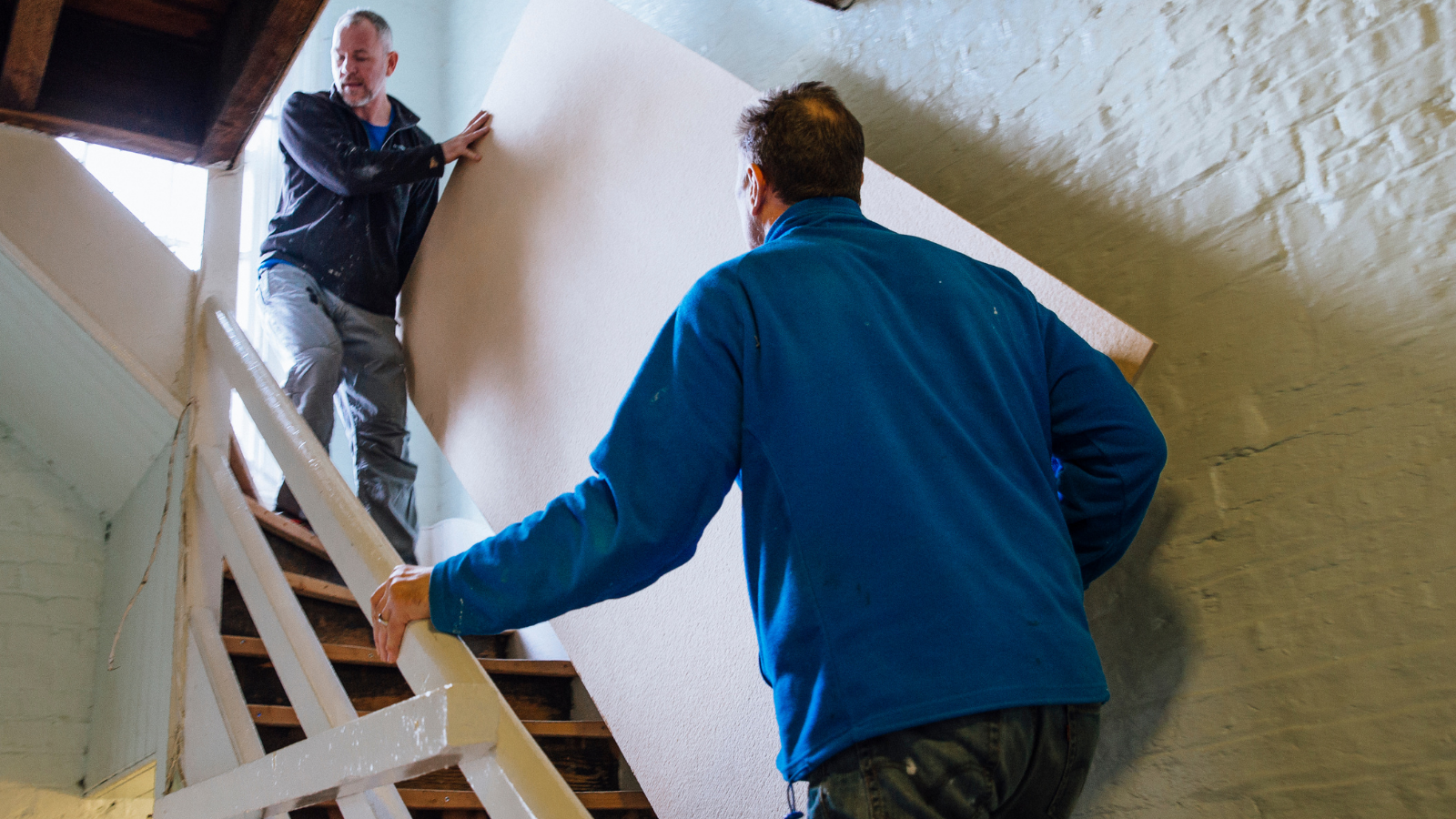Two men are carefully moving a wooden furniture item up a tight staircase. 