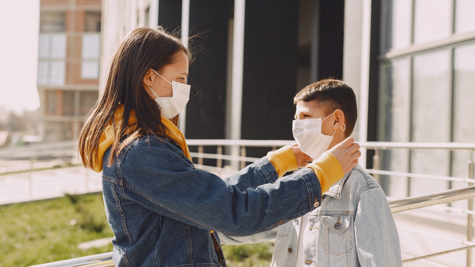 A woman in a yellow jacket is wearing a face mask. She's reaching out and putting a face mask on a young boy in a yellow sweatshirt.