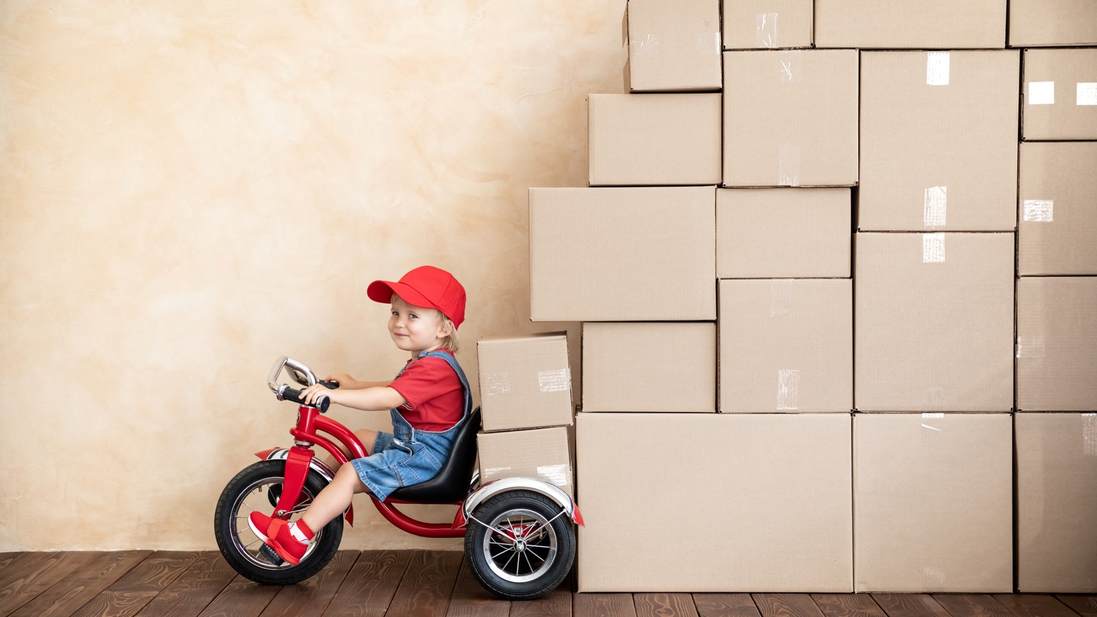 A child riding a tricycle past a vertical stack of packed moving boxes.