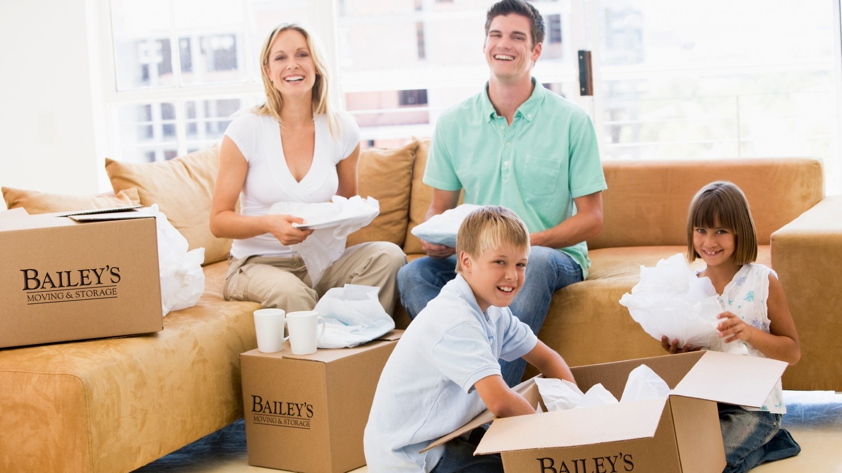 A family sits surrounded by moving boxes, smiling as they unpack. The moving boxes are branded Bailey's Moving & Storage boxes.