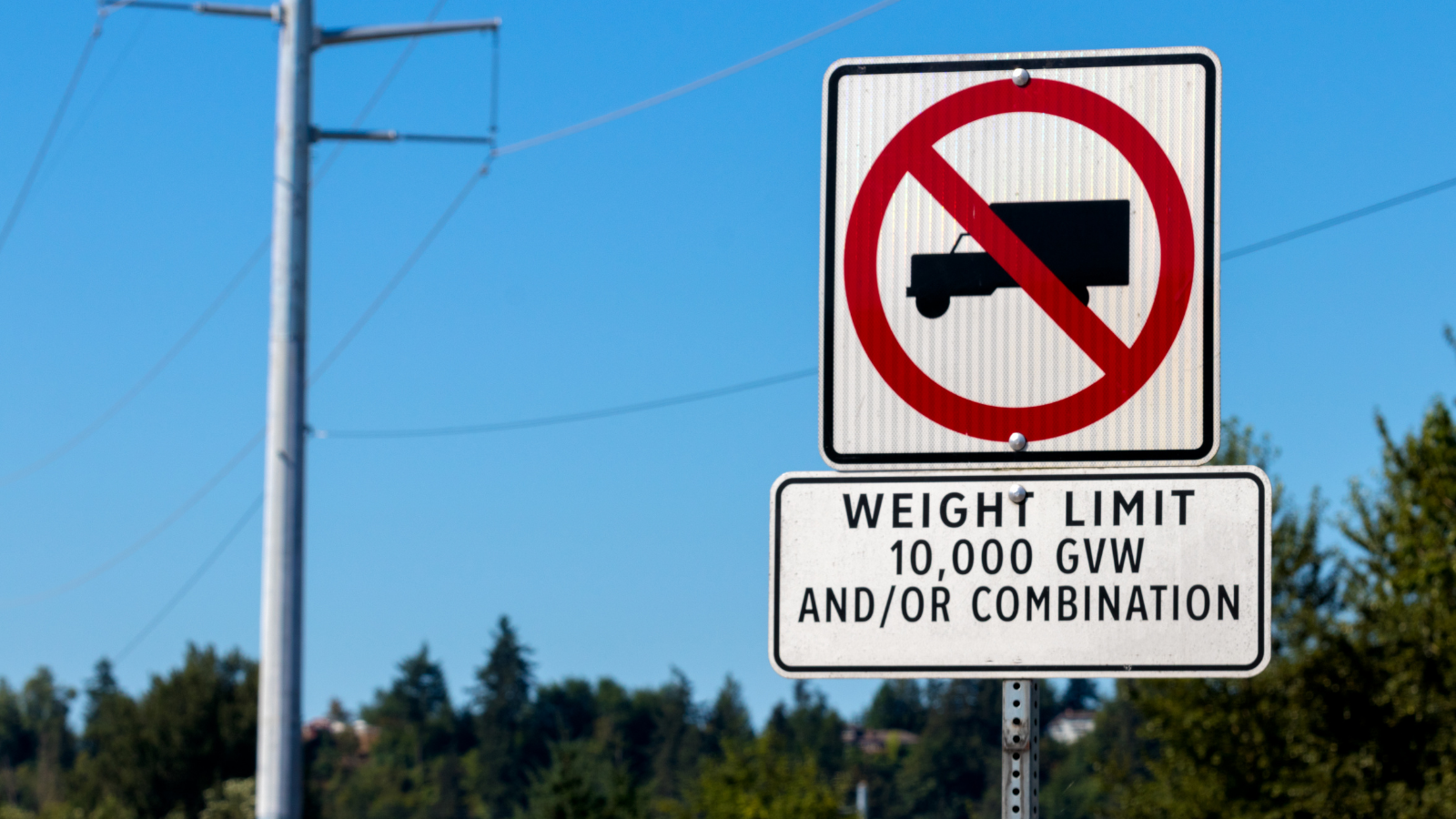 Image of a signpost located on a sunny street that specifies a weight limit for vehicles. The sign has the weight limited at 10,000 GVW and has a graphic of a truck circled in red.