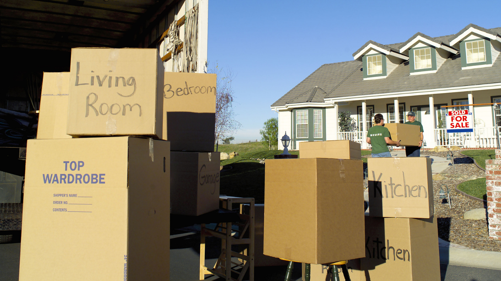 Features moving boxes stacked outside a moving truck in front of a home with a "for sale" sign. 